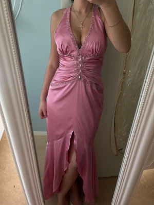 Klänning - Säljer en glansig rosa långklänning köpt i butik med djup v-ringning, smala axelband och dekorativa pärlor framtill. Klänningen har slits framtill och markerad midja. Perfekt för bal, bröllop och fest. Pris dissktuerbart vid snabb affär!  Finns en kort underkjol som slutar precis vid slitsen. Använts endast en gång! Funkar som xs-s  Jag är 167 och klänningen slutar precis vid marken med klack. Pris är disskuterbart vid snabba affärer!