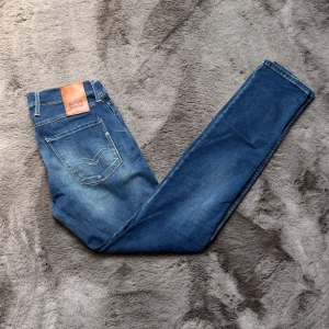 Blå jeans från Replay - Säljer ett par blå Repaly anbass jeans med klassisk femficksdesign och snygga slitningar framtill. Jeansen har en läderpatch med logga bak i midjan och kontrastsömmar längs benen. Perfekta för dig som gillar en modern och avslappnad stil. W30 L32. Priset är ej hugget i sten, hör av dig vid minsta fundering!✨
