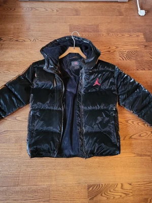 Svart pufferjacka från Jordan - Snygg svart pufferjacka från Jordan med blank finish och röd Jumpman-logga på bröstet. Jackan har huva, dragkedja framtill och två sidofickor. Perfekt för kalla dagar och ger en sportig vibe till din outfit.