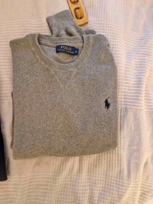 Grå sweatshirt från Polo Ralph Lauren - Klassisk grå sweatshirt från Polo Ralph Lauren med rund halsringning och broderad marinblå logga på bröstet. Tröjan har ribbade muddar vid ärmslut och nederkant, perfekt för en avslappnad och stilren look.