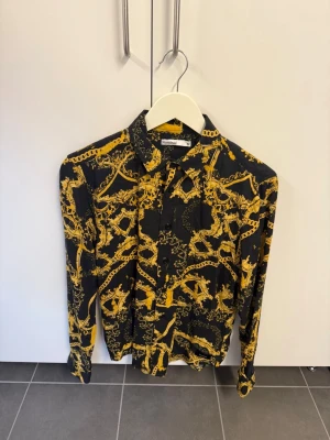 Svart skjorta med gult mönster Nominal, stl S - Snygg svart skjorta från Nominal i storlek S med unikt gult mönster som ger en riktigt cool look. Skjortan har långa ärmar, klassisk krage och knäppning framtill. Perfekt för dig som vill sticka ut med din stil.