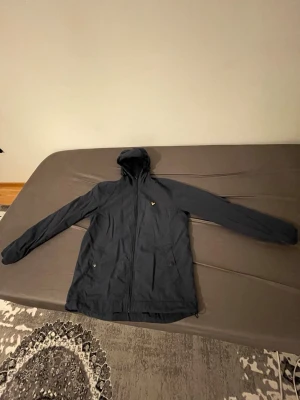 Lyle scott mörkblå jacka - Snygg mörkblå vindjacka från Lyle & Scott med huva och dragkedja framtill. Jackan har två sidofickor med dragkedja och den klassiska gula loggan på bröstet. Tillverkad i lätt och smidigt material som passar perfekt för blåsiga dagar.