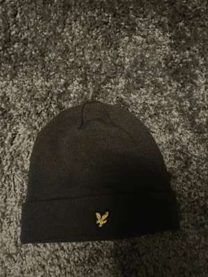 Svart mössa från Lyle & Scott - Säljer en svart ribbstickad mössa från Lyle & Scott med uppvikt kant och den klassiska gula örnloggan framtill. Mössan är i mjukt material och har en clean, stilren look som passar till det mesta.