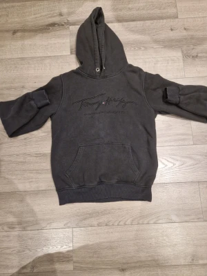 Mörkgrå hoodie från Tommy Hilfiger - Snygg mörkgrå hoodie från Tommy Hilfiger med broderad logga på bröstet. Klassisk känguruficka framtill och huva med dragsko. Tillverkad i mjuk bomullsmix, perfekt för en avslappnad stil.