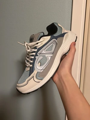 Dior B30 ljusblåa - Säljer ett par Dior B30 sneakers i blått, beige och vitt med coola detaljer och reflekterande inslag. Ovandelen är i mesh och läder med tydlig Dior-logga på sidan. Skorna har vita snören och en chunky sula som ger en modern vibe. Skick: 9/10, aldrig använda. 