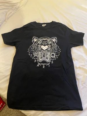 Svart Kenzo t-shirt med tigertryck - Snygg svart t-shirt från Kenzo med det ikoniska tigerhuvudet och texten 'Kenzo Paris' tryckt i vitt på bröstet. Klassisk rund hals och korta ärmar. Perfekt för dig som gillar streetwear och vill sticka ut med ett trendigt motiv. Köpt för 829 använt den max 10 gånger 