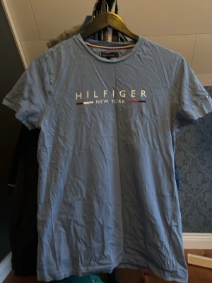 Blå Hilfiger t-shirt med tryck - Snygg blå t-shirt från Tommy Hilfiger med klassiskt tryck 'HILFIGER NEW YORK' på bröstet i vitt,  och rött. T-shirten har rund hals och korta ärmar. Perfekt för en avslappnad och stilren look.