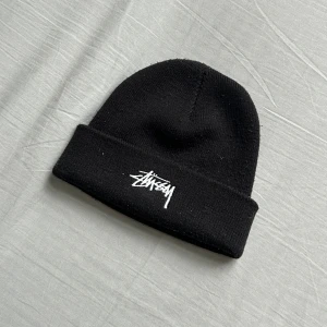 Svart Stüssy mössa  - Säljer en svart mössa från Stüssy med uppvikt kant och vit broderad logga framtill. Klassisk streetwear-look och mjukt stickat material som håller dig varm. Perfekt accessoar för dig som gillar enkel men snygg stil.