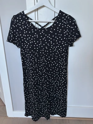 Svart kortklänning med vita prickar - Svart kortklänning med vita prickar och snygg snörning i ryggen. Klänningen har korta ärmar och en avslappnad passform. Perfekt för dig som gillar en enkel men ändå trendig stil.