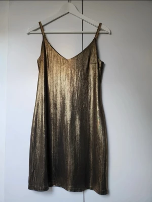 Guldig glittrig klänning med smala band - Säljer en snygg guldig klänning med smala axelband och v-ringning. Klänningen har en glittrig finish och rak passform som ger en cool och trendig look. Perfekt för dig som vill sticka ut med metallic vibes.