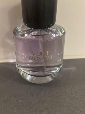 Zara Violet Blossom parfym - Vacker parfymflaska från Zara med namnet Violet Blossom. Flaskan är rund och transparent med en svagt lila tonad vätska och svart kork. Perfekt för dig som gillar blommiga och eleganta dofter. Flaskan är tillverkad i glas och har stilren design.