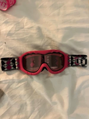 Rosa skidglasögon från Bliz för barn - Säljer ett par rosa skidglasögon från Bliz med rutigt mönster i svart, vitt och rosa på bandet. Glasögonen har tonade linser och ett elastiskt band med Bliz-logga. Perfekt accessoar för vinterns äventyr i backen. Till yngre barn!!