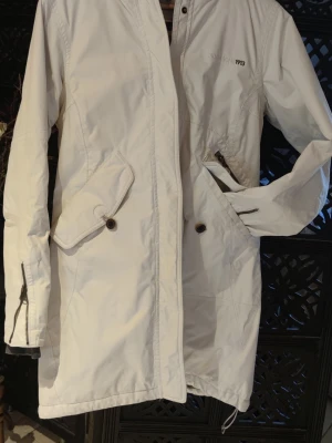 Vit parkas från Didriksons MP13 - Snygg vit parkas från Didriksons MP13 med dragkedja och knappar framtill. Jackan har två stora fickor med lock och knapp, samt en diskret logga på bröstet. Lång modell med justerbar nederkant och muddar vid ärmslut. Perfekt för kyliga dagar.