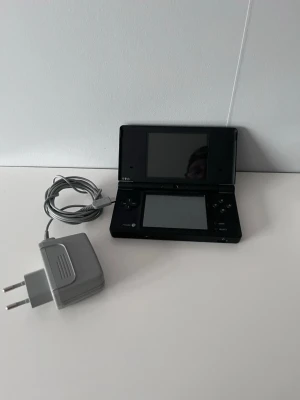 Nintendo DSi svart med spel och laddare - Säljer en svart Nintendo DSi med laddare och sex spel: Pokémon Pearl, Super Mario Bros, FIFA 10, Catz, Imagine: Babyz och Petz. Konsolen har dubbla skärmar, styrkors och knappar. Perfekt för dig som gillar portabel gaming och klassiska DS-spel.