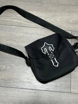 Svart axelväska från Trapstar - Säljer en svart axelväska från Trapstar med justerbar axelrem där loggan är invävd. Väskan har ett stort lock med metallspänne i form av en sköld och dragkedjefack under locket. Materialet är slitstark nylon med meshdetaljer på framsidan.