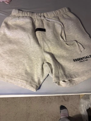 Grå Essentials Fear of God shorts - Säljer ett par ljusgrå shorts från Essentials Fear of God. De har resår i midjan med dragsko och logga tryckt på vänster ben. Mjukt sweatshirtmaterial som är perfekt för chill dagar. Klassisk och avslappnad passform.