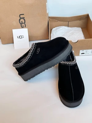Svarta UGG tofflor med platåsula - Säljer ett par svarta UGG tofflor i mocka med kraftig platåsula och dekorativt mönster i vitt runt öppningen. Tofflorna har UGG-logga på sidan och är fodrade för extra komfort. Perfekta för dig som vill ha både stil och mysfaktor.