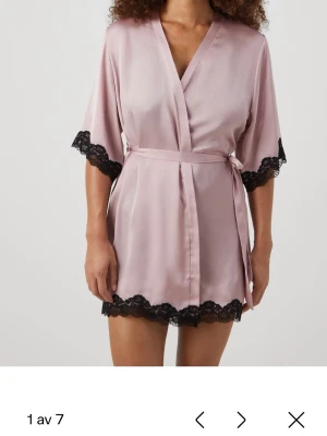 Ljusrosa satinmorgonrock med spets - Kort morgonrock i ljusrosa satin med svarta spetsdetaljer vid ärmslut och nederkant. Knytskärp i midjan och trekvartsärmar ger en avslappnad och elegant look. Perfekt för mysiga hemmakvällar.