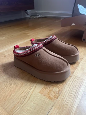 Bruna UGG tofflor med röd kant - Säljer ett par helt nya UGG tazz i storlek 38. Helt oanvända. 