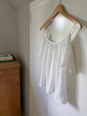 Vit off shoulder topp med volang - Superfin vit offshoulder blus med tunna axelband och vida, luftiga ärmar. Tyget är lätt och har en snygg fallande effekt. Perfekt för en fräsch och stilren look. Passar dig som gillar en luftig och avslappnad vibe. Var tvungen att klippa av lappen då den kliade. Storlek S/M
