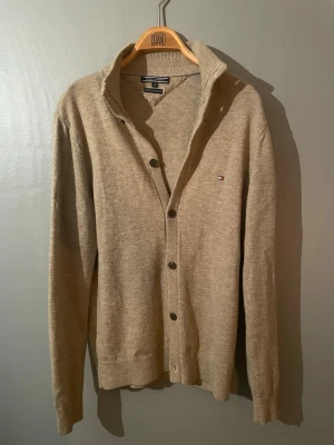 Beige kofta från Tommy Hilfiger - Snygg beige kofta från Tommy Hilfiger med diskret logotyp på bröstet. Koftan har knappar framtill, ribbade muddar och krage. Materialet är mjukt och stickat, perfekt för lager-på-lager. Enkel och stilren design som passar till många outfits. Storlek M