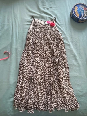 Leopardmönstrad sommarklänning - Snygg ärmlös klänning med leopardmönster i brunt och vitt. Klänningen har smala axelband och en lös, luftig passform som passar perfekt för varma dagar. Dekorerad med en rosa blomdetalj vid axelbandet för en extra touch.