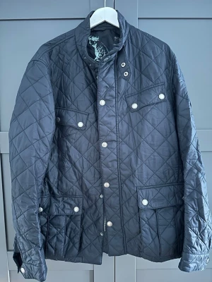 Svart quiltad jacka med fickor - Snygg svart quiltad jacka med fyra framfickor och tryckknappar. Jackan har hög krage och dragkedja samt knappar framtill. Klassisk design med rutmönster och detaljer vid ärmslut. Perfekt för dig som gillar stilrena ytterplagg. OBS passformen är mer lik XL och kommer sitta något tajt om du har XXL 