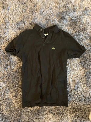 Svart pikétröja från Lacoste - Klassisk svart pikétröja från Lacoste med korta ärmar och krage. Tröjan har knappar vid halsen och den ikoniska gröna krokodilloggan på bröstet. Perfekt till jeans eller shorts för en clean och sportig look.