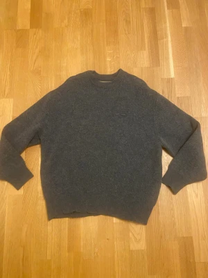 Grå Lacoste Stickad Tröja - Säljer en riktigt snygg och stilren stickad tröja från Lacoste i en mörkgrå melerad färg. Tröjan har den klassiska krokodilloggan broderad på bröstet i en diskret matchande färg (ton-i-ton), vilket ger ett exklusivt intryck. Tröjan är oanvänd utan prislapp, 10/10 skick. Storlek M.