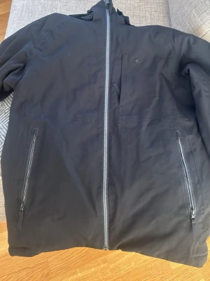 Svart vindjacka från Everest - Snygg svart vindjacka från Everest med huva och hel dragkedja framtill. Jackan har två sidofickor med dragkedja och en bröstficka. Tillverkad i ett slitstarkt material som står emot vind. Perfekt för dig som vill ha en stilren och funktionell jacka.