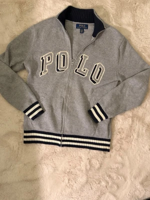 Grå stickad ziptröja Polo Ralph Lauren - Snygg grå stickad ziptröja från Polo Ralph Lauren med stora vita och svarta bokstäver på bröstet. Tröjan har ribbstickade muddar och nederkant med svarta och vita ränder. Passar 160-170