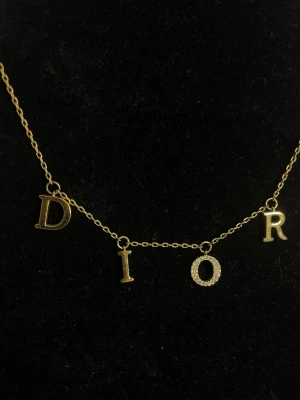 Guldigt Dior halsband med bokstäver - Snyggt halsband i guldfärgad metall från Dior med hängande bokstäver som stavar DIOR. Bokstaven O är dekorerad med små gnistrande stenar och en liten stjärna sitter vid kedjans slut. Perfekt accessoar för dig som gillar lyxiga detaljer.