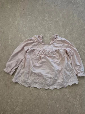 Blus - Söt ljusrosa blus med långärmad design och volangkant nertill. Blusen har broderade detaljer längst ner och en liten volang vid halsen. Knäppning bak i nacken med knapp för enkel påklädning. Perfekt för en romantisk och feminin look.