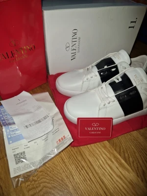 Valentino Garavani vita sneakers - Snygga vita sneakers från Valentino Garavani med en bred svart remsa över sidan. Skorna har klassisk låg profil, vita skosnören och diskret logga på plösen. Tillverkade i slätt läder för en clean och exklusiv look.