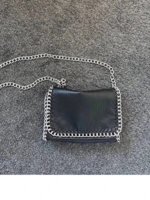 Svart axelväska från tiamo - Snygg svart axelväska i läderimitation med silverfärgad kedja som axelrem och runt kanten. Väskan har en stilren, rektangulär form och lockstängning. Perfekt för dig som gillar trendiga accessoarer med lite edge.