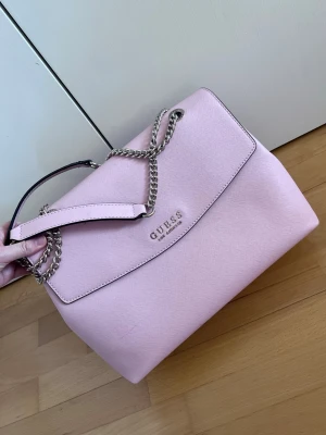 Babyrosa handväska Guess - Supsersnygg babyrosa handväska från Guess. Aldrig använd men har fått ett litet sträck under transport (se bild). 