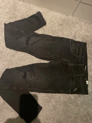Svarta slim fit jeans från Replay - Säljer ett par svarta slim fit jeans från Replay med slitningar vid knäna och klassisk femficksdesign. Jeansen har en snygg tvätt och detaljerade sömmar, samt Replay-logga på bakfickan. Perfekta för en trendig och avslappnad stil.