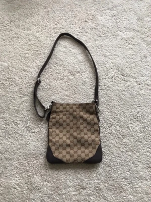 Gucci brun axelväska med monogram - Säljer en snygg brun axelväska från Gucci med klassiskt GG-monogram i beige och brunt. Väskan har justerbar axelrem, detaljer i mörkbrunt läder och dragkedja upptill. Insidan har ett fack med dragkedja för småsaker. Perfekt för dig som gillar stilrena designer.