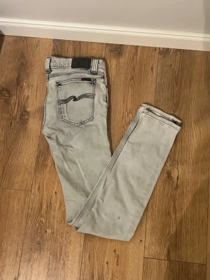 Ljusgrå jeans från Nudie - ( kan göra byten mot andra kläder)Säljer ett par ljusgrå jeans från Nudie med klassisk femficksdesign och svarta kontrastsömmar. Jeansen har en svart patch bak i midjan och Nudies ikoniska broderi på bakfickan. Perfekta för en avslappnad och trendig look.