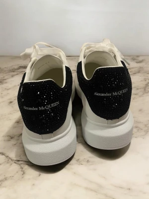 Alexander McQueen Limited edition black stars - Stilrena vita sneakers från Alexander McQueen med svart hälparti täckt av svarta glittrande stenar och logga. Skorna har tjock vit sula, vita skosnören och är tillverkade i slätt läder. Perfekta för dig som vill ha en lyxig och trendig look.