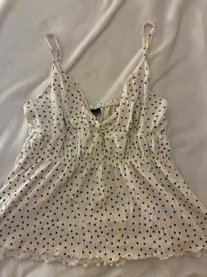 Prickig vit linnetopp med smock - Söt vit linnetopp med svarta prickar och smockad midja. Toppen har tunna axelband och v-ringad hals, vilket ger en luftig och avslappnad känsla. Perfekt för varma dagar och enkel att matcha med jeans eller kjol.