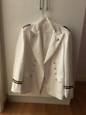 Vit kavaj med guldknappar - Stilren vit kavaj med dubbla rader guldknappar och svarta axel- och ärmdetaljer med guldrand. Klassisk krage och två fickor framtill. Perfekt för dig som vill sticka ut med en unik och elegant look.Navy. Kapten kavaj, storlek XXXL 