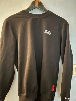 Svart sweatshirt från Hugo Boss - Snygg svart sweatshirt från Hugo Boss med rund halsringning och diskret logga på bröstet. Tröjan har ribbade muddar vid ärmslut och nederkant samt en röd detalj vid sidan. Perfekt för en clean och stilren look. Först till kvarn som gäller, passar den som har xs/S