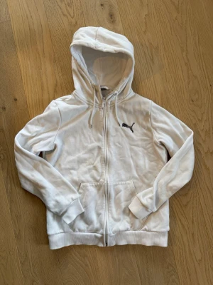 Vit hoodie med dragkedja från Puma - Snygg vit hoodie från Puma med dragkedja framtill och svart Puma-logga på bröstet. Utrustad med huva och dragsnören samt två fickor framtill. Perfekt för en avslappnad och sportig stil. Stl M för Junior (XS herr)