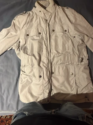 Vit vindjacka från Hugo Boss - Stilren vit vindjacka från Hugo Boss med hög krage, flera fickor med knappar och dragkedja framtill. Jackan har en modern look och är tillverkad i ett lätt syntetmaterial som passar perfekt för blåsiga dagar.