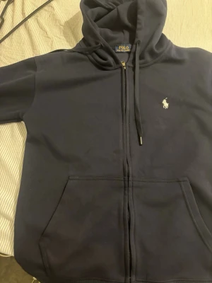 Mörkblå hoodie från Polo Ralph Lauren - Snygg mörkblå hoodie från Polo Ralph Lauren med dragkedja framtill och klassisk vit logga på bröstet. Hoodien har huva med dragsko och en stor magficka. Tillverkad i mjukt bomullsmaterial som är skönt att bära.