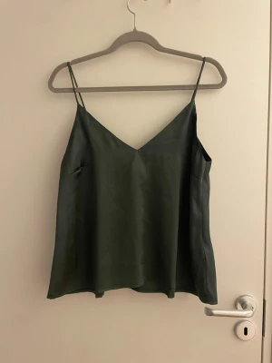 Mörkgrön linnetopp med smala band - Snygg mörkgrön linnetopp med tunna axelband och v-ringad hals. Toppen har en lös passform och är tillverkad i ett glansigt material som ger en lyxig känsla. Perfekt att styla med jeans eller kjol för en trendig look.