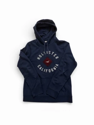 Mörkblå hoodie från Hollister - Mörkblå hoodie från Hollister med broderad logga och texten 'Hollister California' på bröstet. Klassisk känguruficka framtill och justerbar huva med dragsko. Mjuk bomullsblandning som känns skön mot huden. Perfekt för en avslappnad och trendig look.