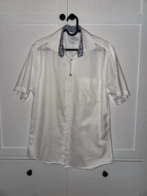 Vit kortärmad skjorta från Erla - Vit kortärmad skjorta från Erla med klassisk krage och diskret blommigt mönster på insidan av kragen. Skjortan har en bröstficka och knäppning framtill. Tillverkad i ett mjukt och lätt tyg, perfekt för varma dagar.