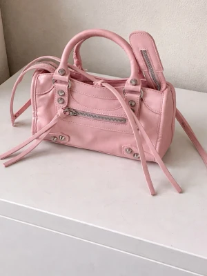 Rosa City Bag 15x24 cm – lyxig stil, Y2K - Vacker rosa miniväska i en lyxdesigner-inspirerad citybag-stil. Den har en mycket trendig Y2K-look och lyfter vilken outfit som helst, samtidigt som den är praktisk för både vardag och utekvällar. Väskan har aldrig använts och är därför i utmärkt, som-nytt skick. Den rymmer perfekt telefon, kort, nycklar och andra dagliga nödvändigheter och kan bäras i handen eller med remmen som axel- eller crossbodyväska. Den mjuka rosa färgen och ikoniska formen gör den till en mycket eftertraktad